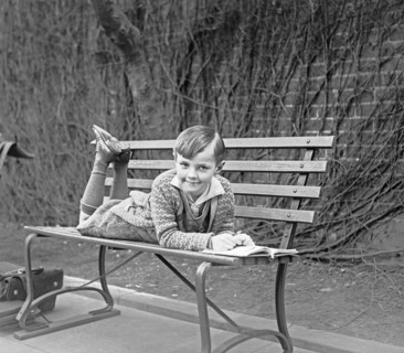 Child lying on outdoor bench with an open book ca. between 1909 and 1923