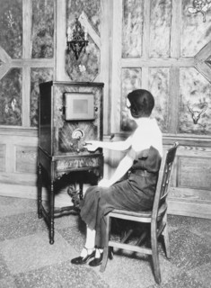 Woman, full-length, seated, facing left, turning dial of a console radio ca. 1909