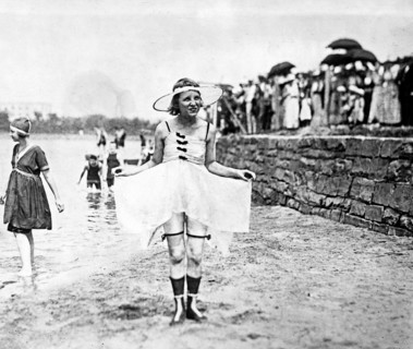 Young woman posing in bathing suit with other bathers and spectators in background ca. 1909