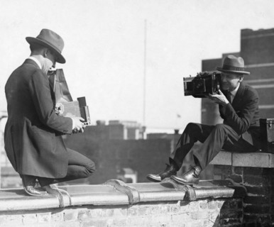 Two photographers taking each other's picture with hand-held cameras while perched on a roof ca. 1909