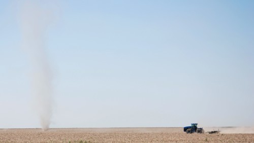 Farmer plowing a field on the Eastern Colorado plains, tractor facing left and driving toward a natural phenomena called a dust devil