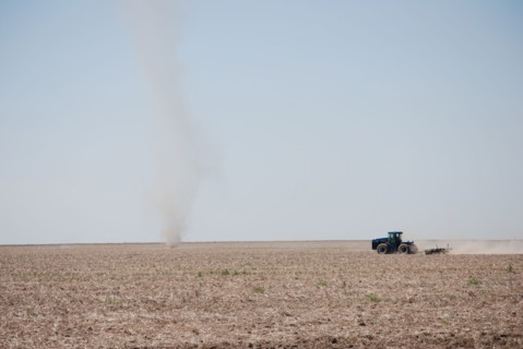 Farmer plowing a field on the Eastern Colorado plains, tractor facing left and driving toward a natural phenomena called a dust devil