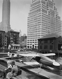 Two shoe-shine boys sit on edge of pier overlooking skyport, where seaplanes are moored; Manhattan office buildings beyond ca. 1936