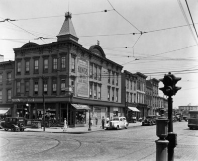 Clapboard buildings, cobblestone streets with trolley tracks, cars, including a taxi and an entrance to the subway on the corner. Graham and Metropolitan Avenues, Brooklyn. ca. 1937