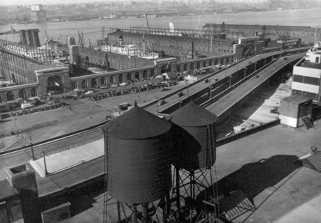 Eight cars visible on Westside highway, beyond water towers in foreground, piers have entrances on street, ships visible at dock ca. 1937