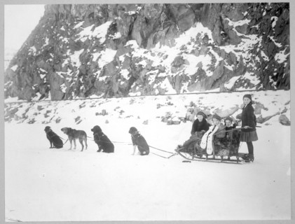 Dog team harnessed to a sled holding four girls, in front of a snow-covered cliff. 1900-1917 Canada