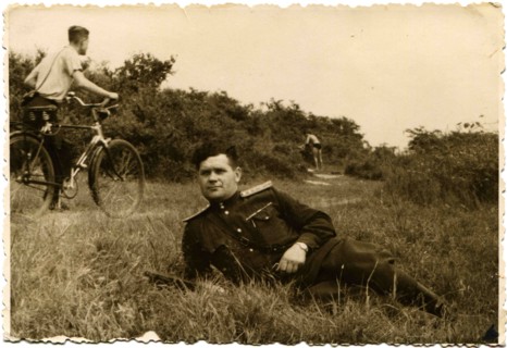 Soviet Army captain lying in the grass on the banks of the Elbe River in the background a man riding a bicycle on a footpath.