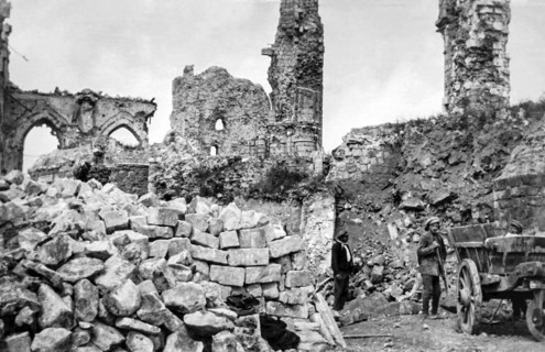 Ruins of buildings and a pile of rubble and stone blocks with workers standing by a wooden cart.