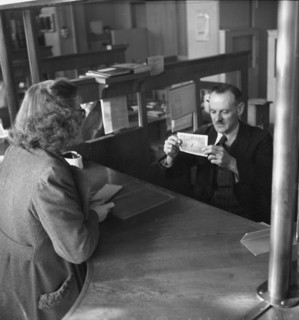 Client at post office counter; post office official checking banknote for its authenticity;  1943.