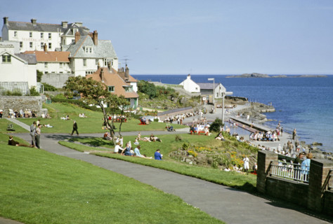 People sittng on grass by the coast enjoying sunshine at the seaside resort of Portrush, County