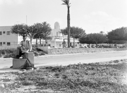 Young man sitting on low wall of water fountain on street.