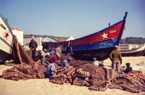 Fishermen repairing nets on beach by traditional fishing boats.