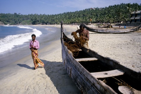 INDIA, Kerala, Kovalam, Fisherman tending to his boat moored on the sandy beach with passing man in
