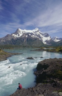 CHILE, Patagonia, Torres Del Paine, Man sitting on the riverbank beside rapids below the distant