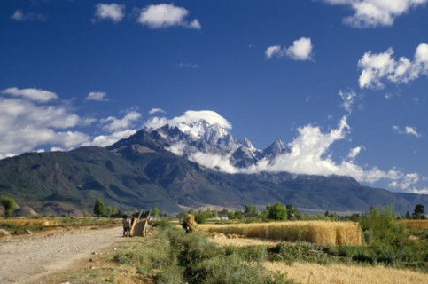 CHINA, Yunnan, Lijiang, Yalong Mountain with cart and people harvesting wheat in the foreground