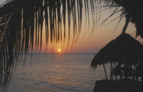 WEST INDIES, Jamaica, Negril, Ricks Cafe at sunset through coconut palm tree with tourist sitting