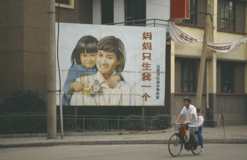 CHINA, Shandong, Jinan, Man with child passenger cycling past birth control poster with image of