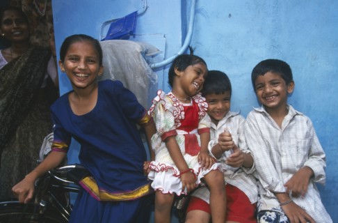 INDIA, Maharashtra, Mumbai, Four smiling children leaning against bicycle with blue wall behind.