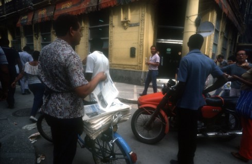 CUBA, Havana, Newspaper vendor selling goods from a bicycle seat in the street with passing traffic