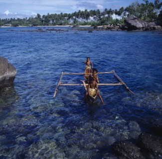 INDONESIA, Irian Jaya, Sorong, "Children in outrigger canoe on clear dark blue sea, rocks,palms "