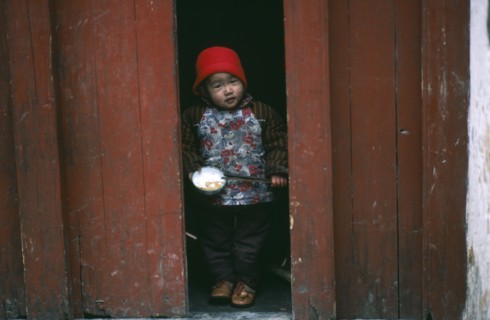 CHINA, Guangxi, Yangshuo, Child holding bowl of rice and chopsticks looking out from narrow doorway