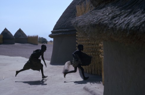 20049468 SUDAN People Children Shilluk children running towards thatched village hut.