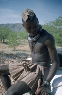 NAMIBIA, Skeleton Coast, Hoarusib Valley, Kaokoland.  Himba man sitting on bumper of safari jeep.