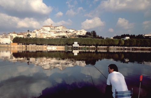 PORTUGAL, Coimbra, Man fishing on the bank of the River Mondego with the University crowning