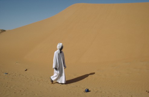 SUDAN, Kordofan Province, El Humra Village, Man in white robe and turban passing huge sand dunes