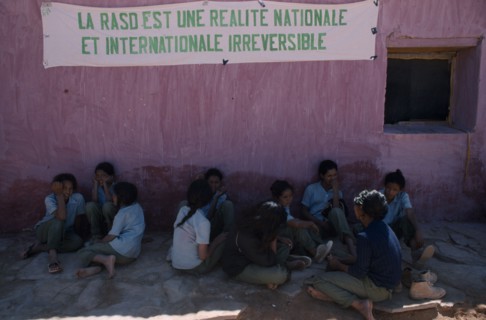 WESTERN SAHARA, SADR, Sahrawi schoolgirls sitting against pink plastered wall under banner with