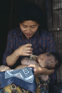 THAILAND, People, Cambodian refugee mother breastfeeding baby while sewing.