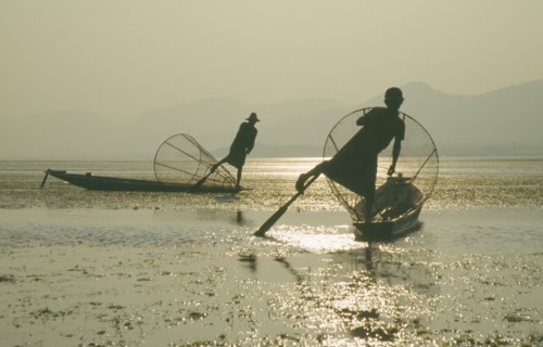 MYANMAR, Shan State, Inle Lake, Silhouettes of two Intha fishermen with Sun’s reflection on water.