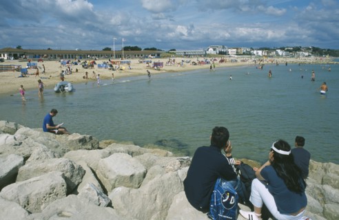 ENGLAND, Dorset, Sandbanks, People sat on rocks looking across water and busy sandy beachfront