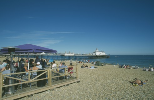 ENGLAND, East Sussex, Eastbourne, Beach Cafe with people sat on chairs under parasols and the pier