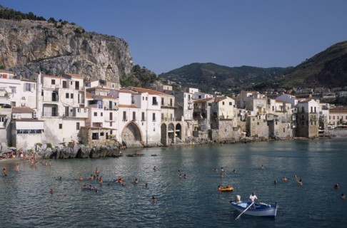 ITALY, Sicily, Palermo, Cefalu. View across people swimming in the sea next to sandy beach with a