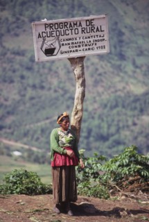 GUATEMALA, Huehuetenango, "Indigenous woman standing below a sign describing a village water
