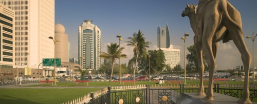 UAE, Dubai, "Camel statue in Al-Ittihad Square, people sat on grass. Palm trees and car park in