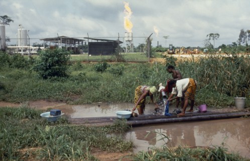 NIGERIA, Environment, Women washing clothes in foreground with oil refinery behind.