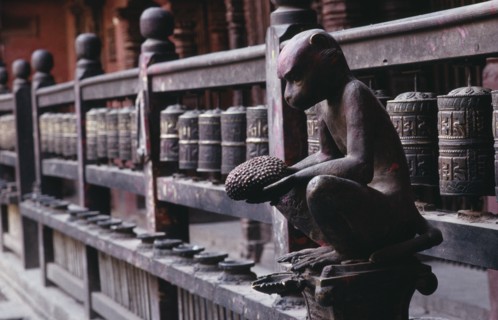 NEPAL, Patan, Carving of monkey holding a jack fruit in front of mane or prayer wheels behind.