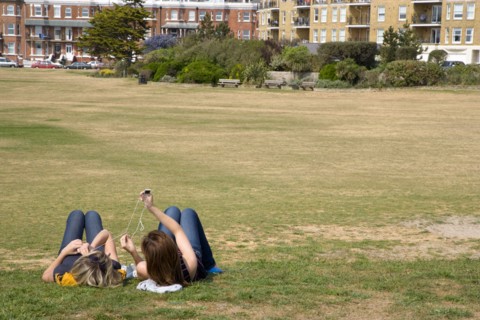 ENGLAND, West Sussex, Littlehampton, Two girls lying on grass by seafront promenade using and