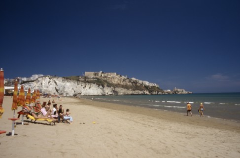 ITALY, Puglia, Foggia, Vieste.  Line of sunbathers on stretch of sandy beach overlooked by castle