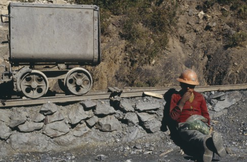 BOLIVIA, Chukiuta, Miner wearing orange hard hat sitting next to mining rail track chewing leaves