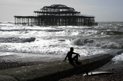 ENGLAND, East Sussex, Brighton, "Seafront, man sat with surf board on goyne in front of the burnt