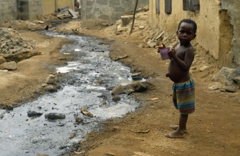 Ghana, Dixcove, Child holding drinking cup standing beside open sewer through street centre.