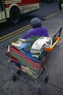 England, East Sussex, Brighton, Homeless lady sitting on a pavement bench beside a road with a bus