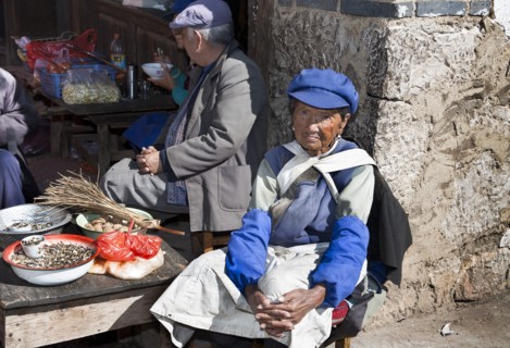Old Naxi woman wearing traditional costume selling goods on market stall in the village near
