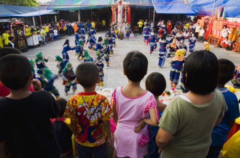 Thai children watch dance troupe at local temple from elevated stage.