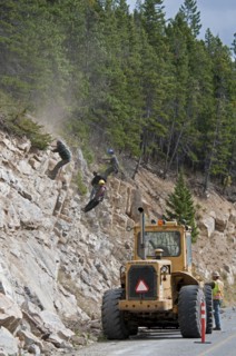 Workers wearing safety helmets on ropes clearing a rock slide and making the rock face safe along the Akamina Parkway Caterpillar tractor on parkway