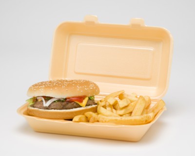 Food, Cooked, Meat, Cheesburger with salad and tomato ketchup in a bun with potato chips inside a polystyrene foam box on a white background.