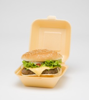Food, Cooked, Meat, cheesburger with salad and tomato ketchup in a bun inside a polystyrene foam box on a white background.